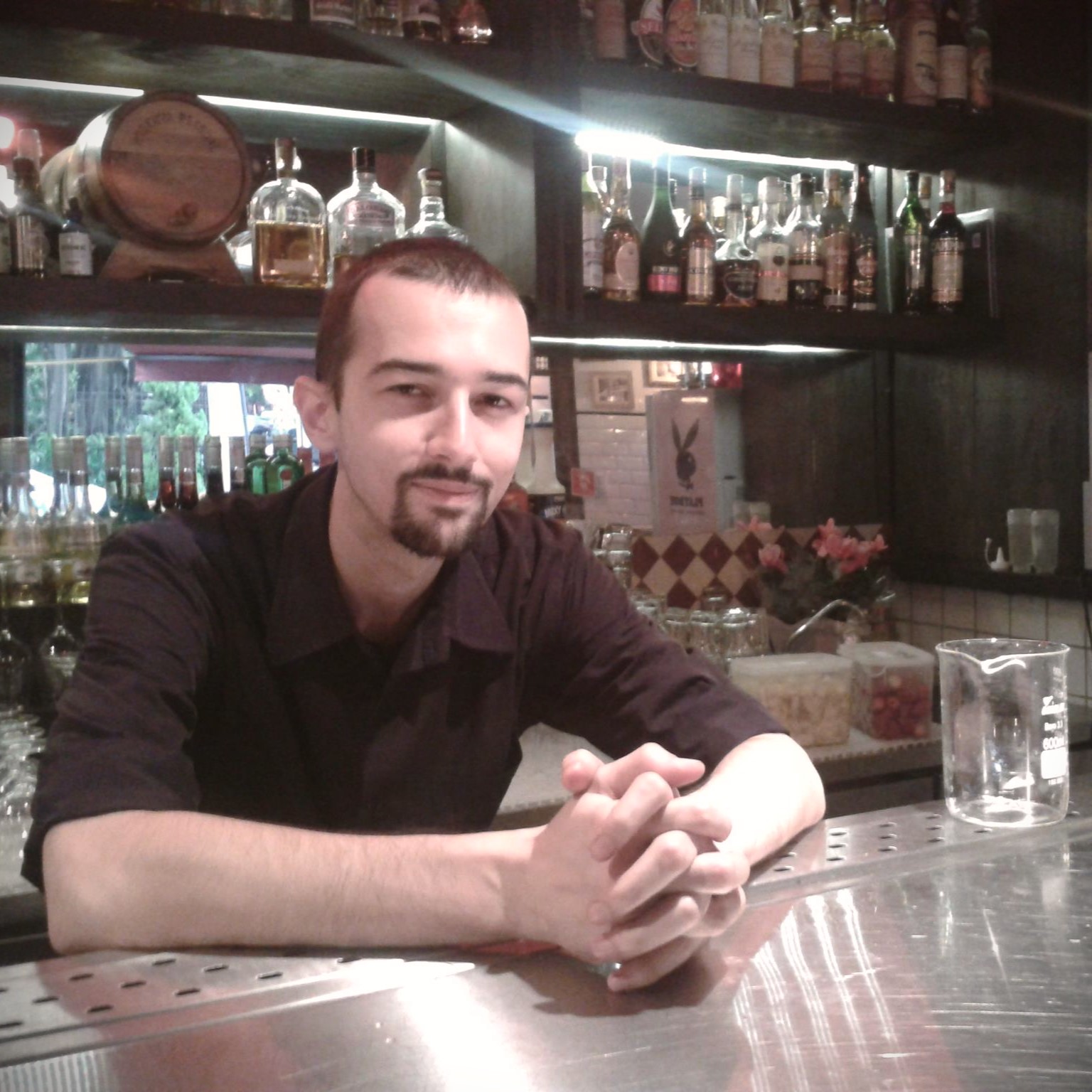 foto do portfólio do Emerson. A imagem mostra um balcão de bar com várias garrafas de bebidas alinhadas em prateleiras ao fundo. Há uma pessoa com cavanhaque, vestindo uma camisa escura e com as mãos entrelaçadas sobre o balcão. O balcão tem um acabamento metálico e há um mixing glass vazio ao lado da pessoa. A iluminação é suave e indireta, criando um ambiente acolhedor.